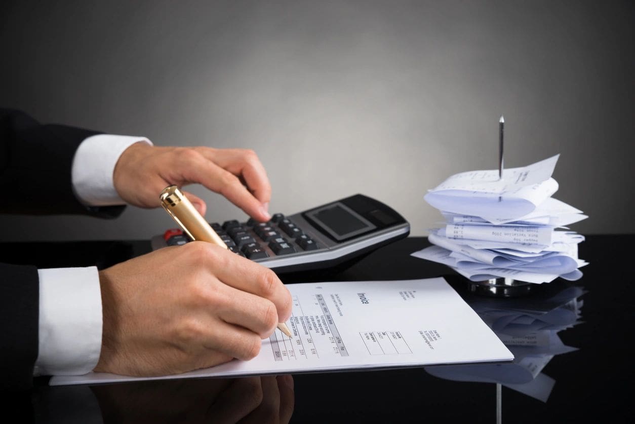 A person calculating expenses and writing on a document with a calculator and stacks of receipts on the desk.