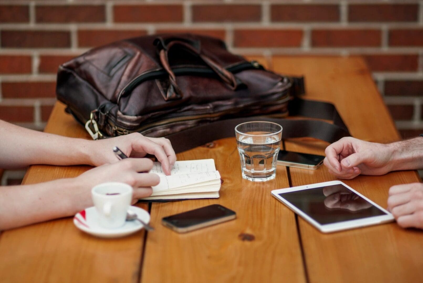 Two people sitting at a wooden table with a cup of coffee, a glass of water, mobile devices, and a leather bag.
