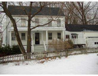 A white two-story house with a front porch, surrounded by a picket fence in a snowy setting.