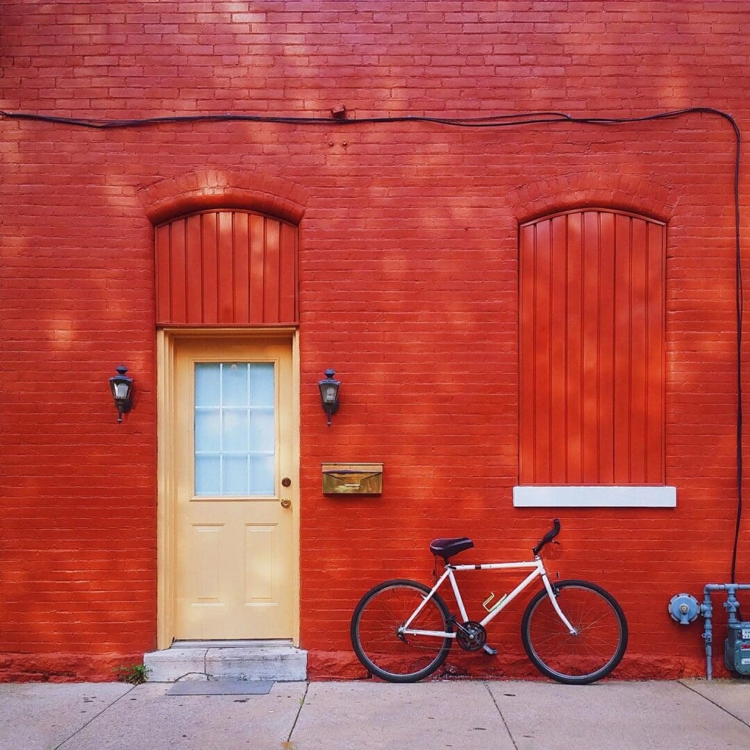 A vibrant red brick wall with a yellow door, two windows covered by red wooden panels, a bicycle parked on the side, and outdoor light fixtures.