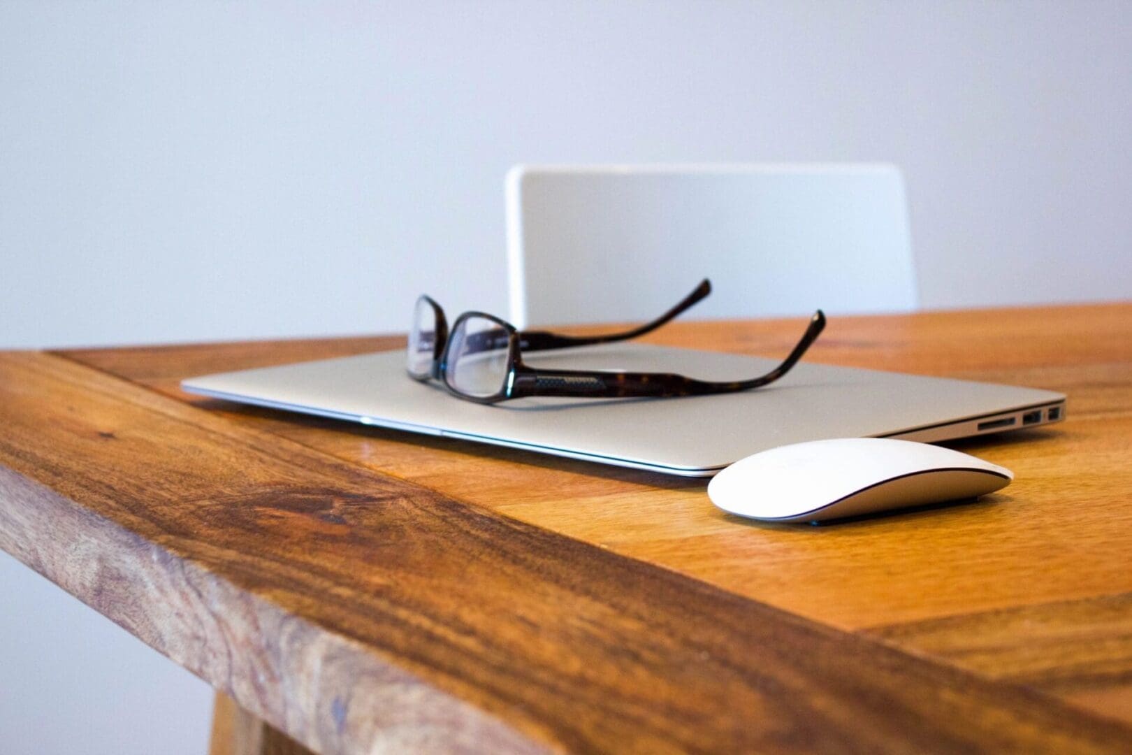 A pair of eyeglasses and a wireless mouse resting on a wooden table beside a closed laptop.