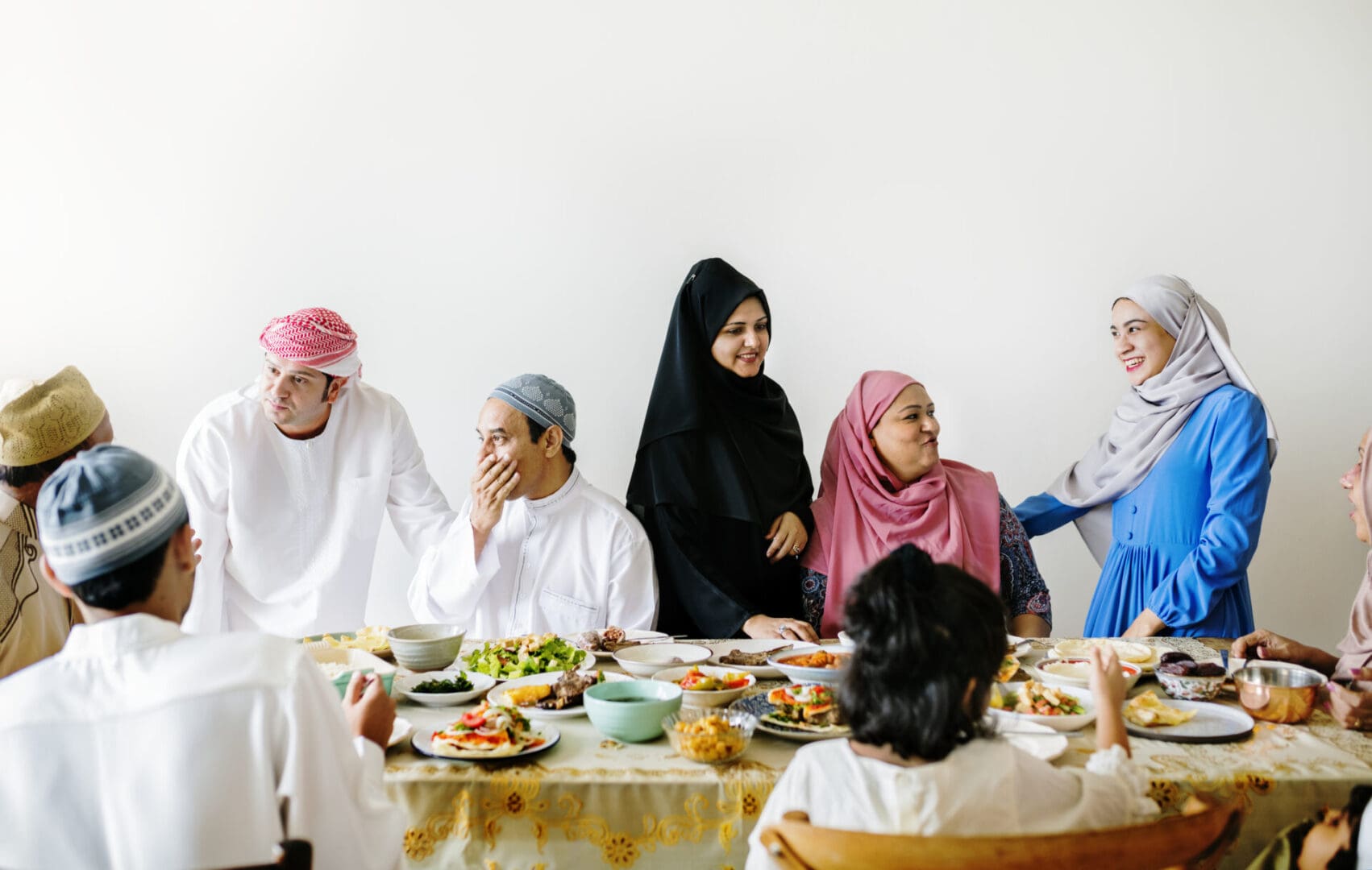 A family gathering for Eid Al-Fitr with members dressed in traditional Middle Eastern attire sharing a meal and conversation around a dining table.