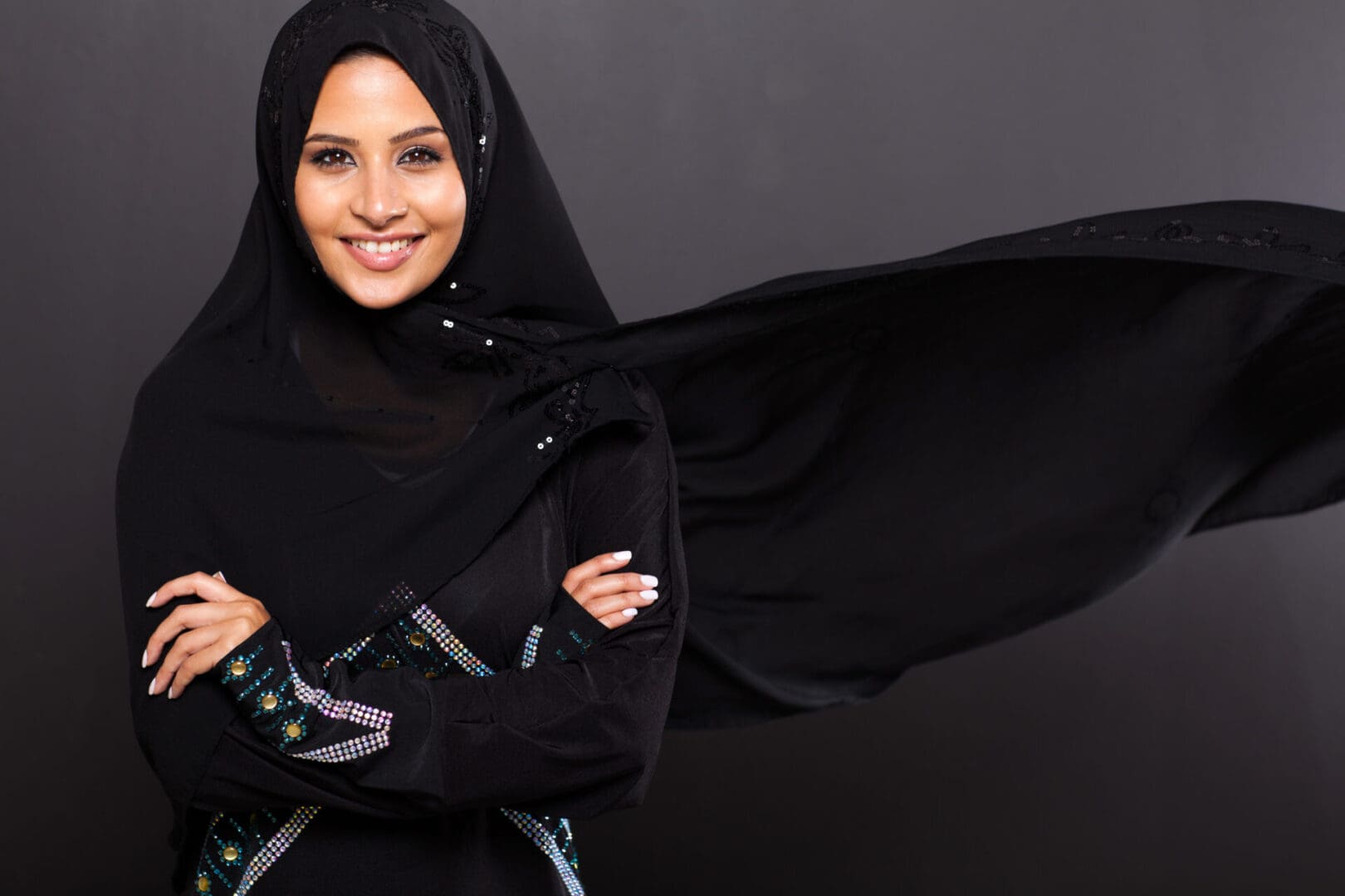 Woman in a decorated black hijab smiling at the camera with her arms crossed.
