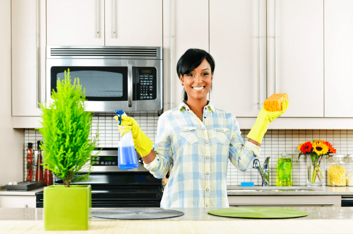 A woman smiling while wearing gloves and holding spring cleaning supplies in a kitchen.