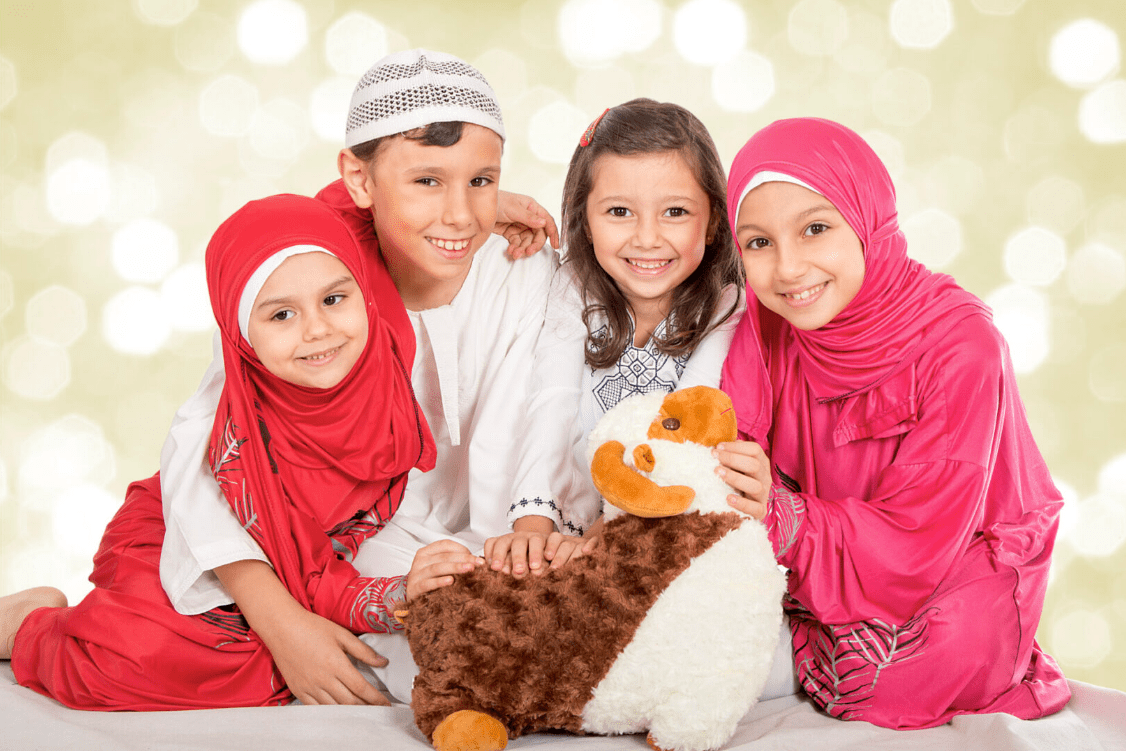 Four children smiling at the camera, with two girls wearing pink hijabs and one boy wearing a white cap on Eid; the youngest girl is holding a plush toy.