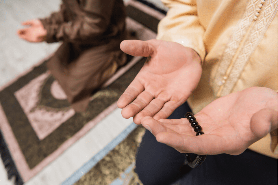 Hands holding prayer beads with another individual sitting in the background on a prayer mat during Ramadan.