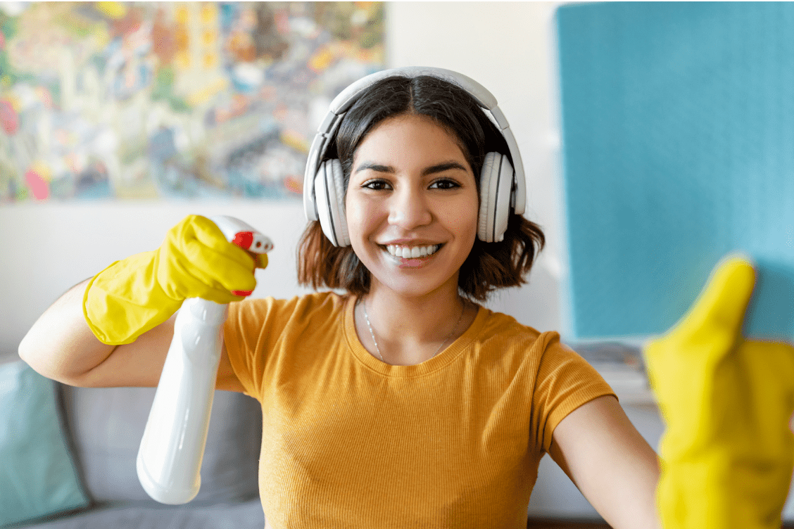 Woman wearing headphones and gloves holding an allergen cleaning spray bottle while smiling at the camera.