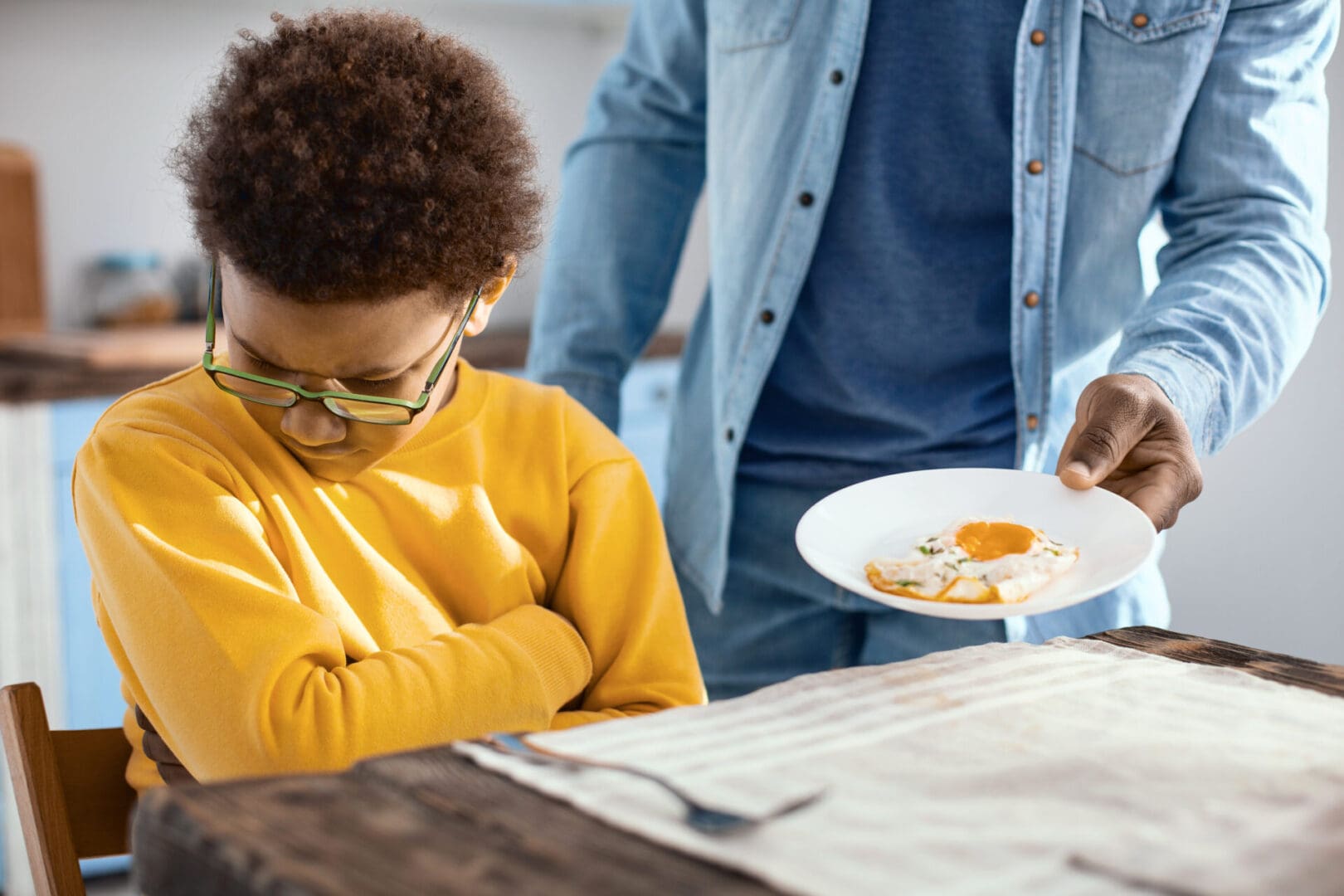 Child, a picky eater, showing disinterest in food while an adult offers a plate with an uneaten meal.