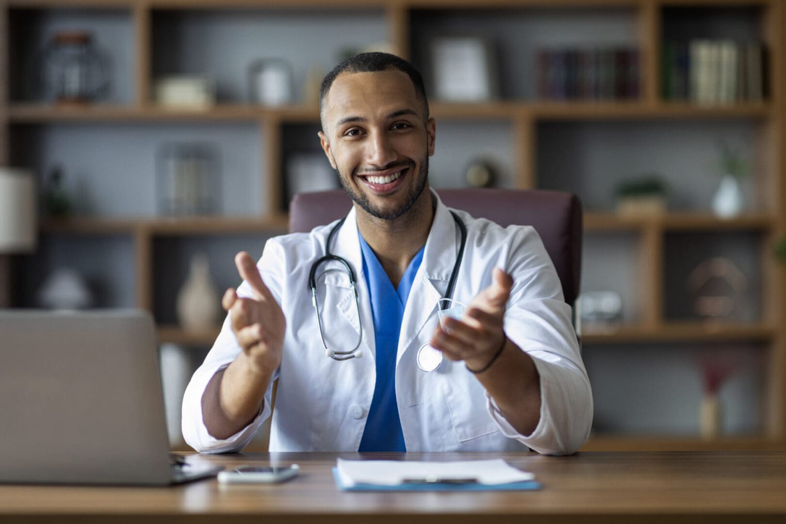 Cheerful african american doctor sitting at workdesk at clinic, gesturing A smiling healthcare professional gesturing invitingly during a consultation in a US residential program.