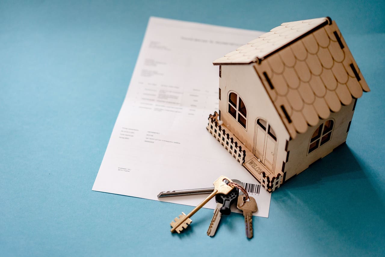 A wooden model house next to a set of keys and a document on a blue background, symbolizing residential programs in Canada.
