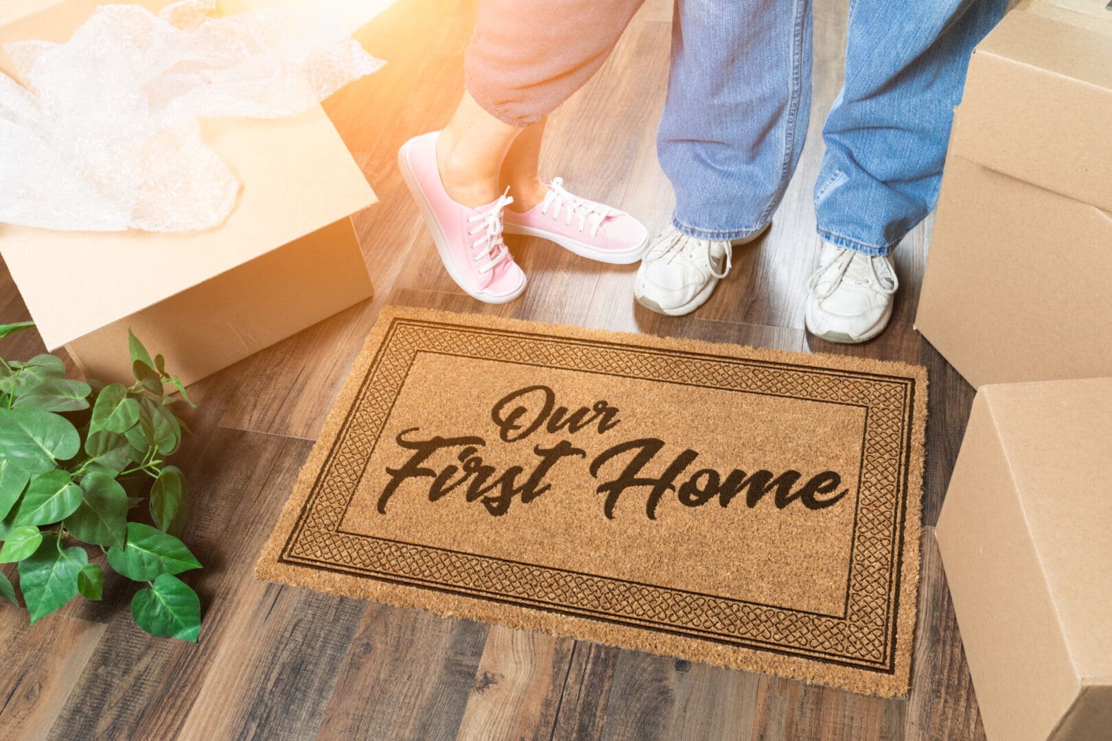 Two first-time home buyers standing next to a "our first home" doormat, surrounded by moving boxes.