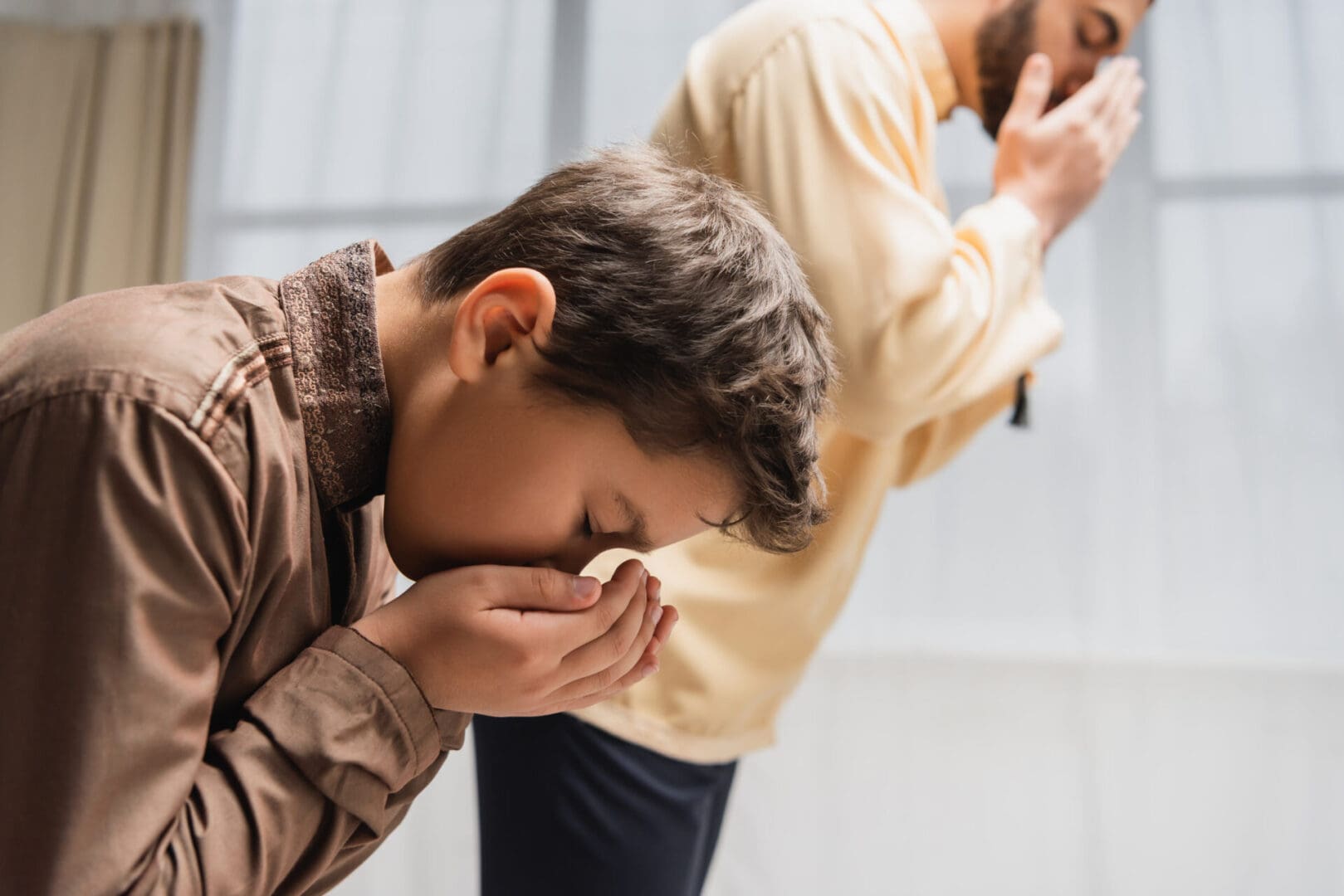 Young boy sneezing into tissue with another person standing in the background during Ramadan.