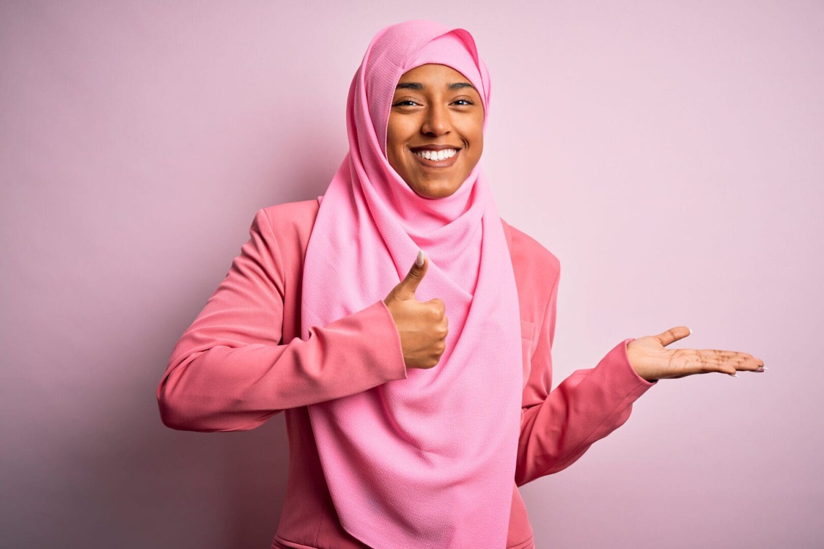 Young African American afro woman wearing muslim hijab over isolated pink background Showing palm hand and doing ok gesture with thumbs up, smiling happy and cheerful