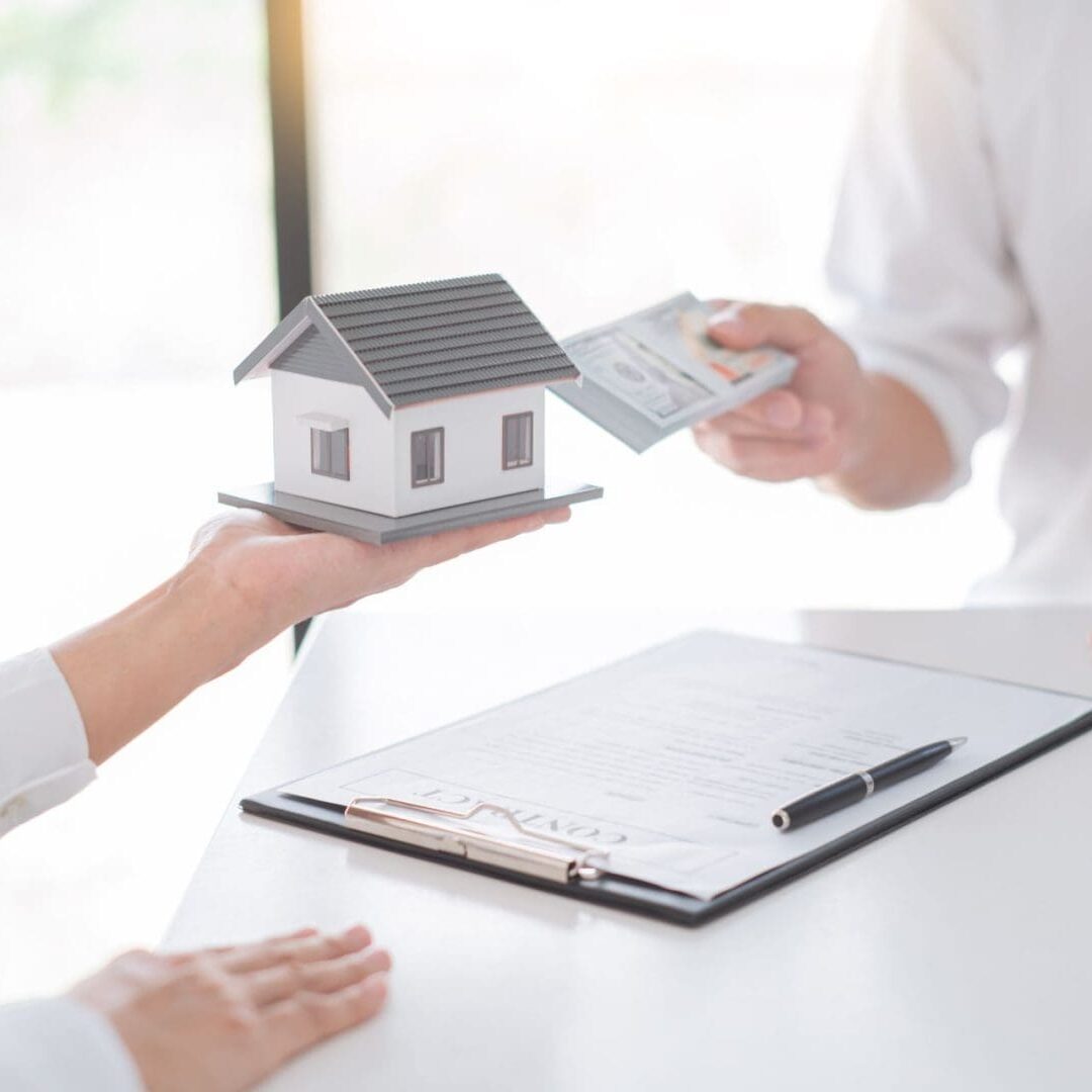 Residential real estate transaction: one person handing over cash in exchange for a house model with contract documents on the table.