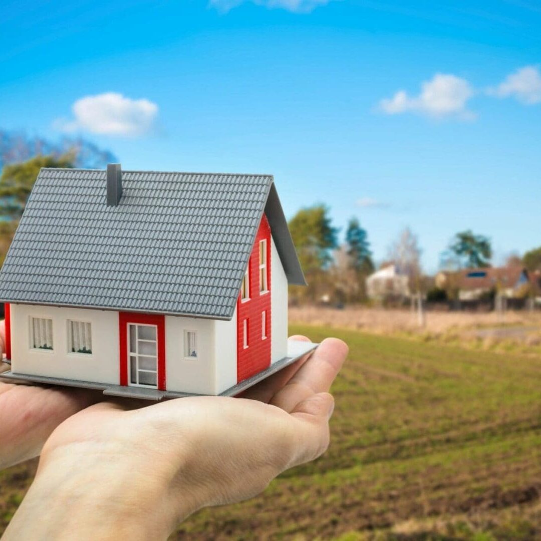 Hand holding a small model of a red and white residential house with a background of a suburban landscape under a blue sky.