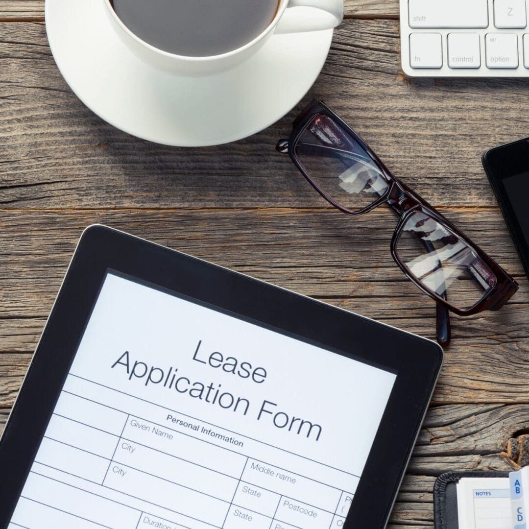 A digital tablet displaying a residential lease application form on a wooden desk, alongside a cup of coffee, eyeglasses, a smartphone, and a computer keyboard.