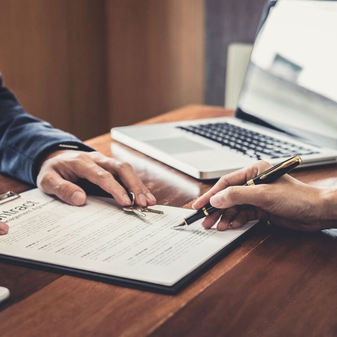 Two individuals in a residential business setting, with one signing a document and the other pointing at a specific section, suggesting a discussion about the content. A laptop and mobile phone are also present on the desk