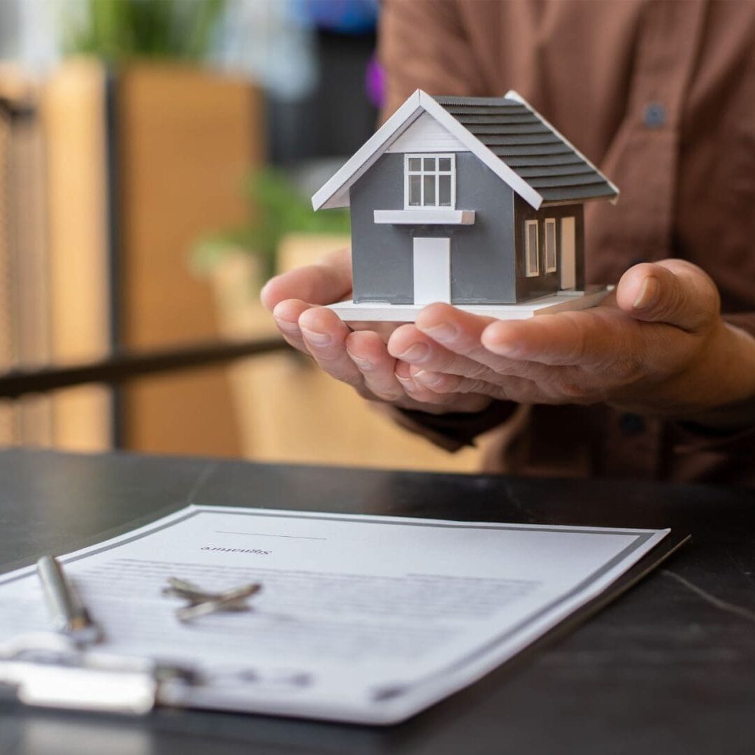 Person holding a small house model with a contract and keys on the table, symbolizing real estate ownership or sale.